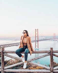 a woman sitting on top of a wooden fence next to the ocean and bridge in the background