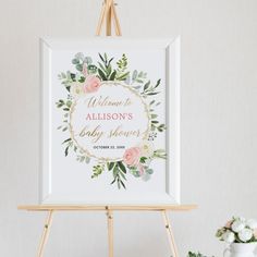 a baby shower sign on an easel next to a vase with flowers and greenery