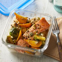 a glass container filled with salmon and vegetables on top of a table next to a fork