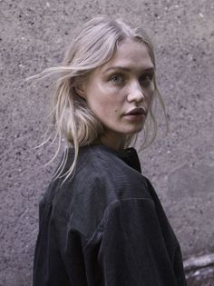 a woman with blonde hair standing in front of a concrete wall and looking at the camera