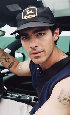 a man with tattoos on his arm sitting in the drivers seat of a car wearing a hat