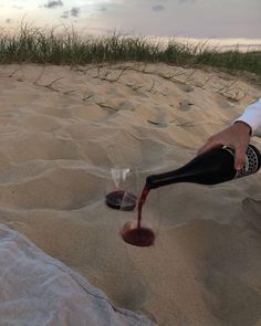 a person pouring wine into a glass on the beach at sunset with grass and sand in the background