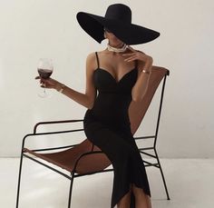 a woman sitting in a chair holding a glass of wine and wearing a black hat