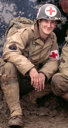 a man in uniform sitting on the ground next to another man wearing a helmet and boots