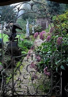 an iron gate in the middle of a garden filled with flowers and statues, surrounded by greenery