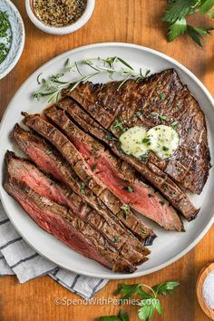 steak on a white plate with herbs and seasoning next to it, sitting on a wooden table