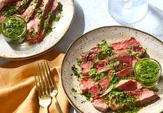 two plates with meat and pesto on them next to a fork, knife and glass of water