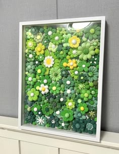 an art piece made out of green and yellow flowers in a white frame on top of a shelf