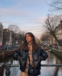 a woman standing next to a body of water wearing a black jacket and plaid scarf