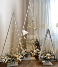 three christmas trees with ornaments and lights on them in front of a curtained window