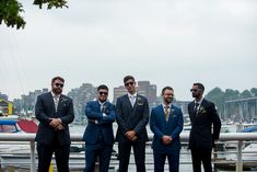 four men in suits and sunglasses standing next to each other on a dock with boats in the background