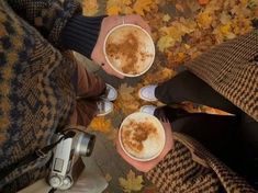 two people holding cups of coffee in their hands with leaves on the ground around them