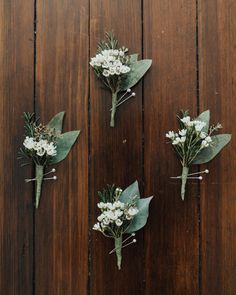 three boutonnieres with white flowers and green leaves on a wooden surface,