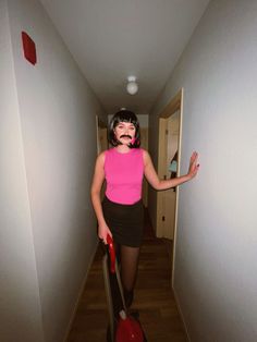 a woman in pink shirt and black skirt with suitcase walking down hall way wearing fake moustache