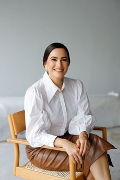 a woman sitting in a chair with her legs crossed and wearing a white shirt and brown leather skirt