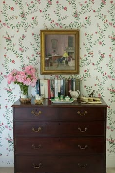 a dresser with flowers and books on it in front of a wallpapered wall