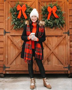 a woman standing in front of a wooden door with wreaths on it and holding a cup