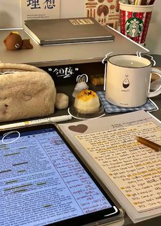 an open book on a desk next to a tablet computer and coffee mug with writing