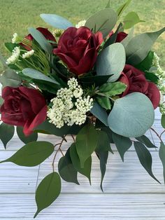 a bouquet of red roses and greenery on a white wooden table with grass in the background