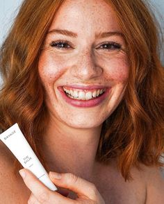 a woman with freckled red hair smiles while holding a tube of sunscreen