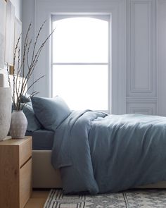 a bed with blue sheets and pillows in front of a large window next to a dresser