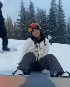 a woman sitting in the snow on top of a snowboard with trees behind her