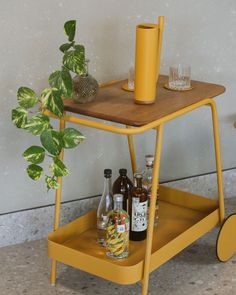 a yellow cart with drinks on it next to a potted plant and two glasses