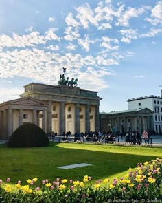 people are walking around in front of a building with statues and flowers on the lawn