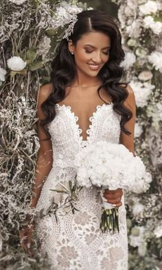 a woman standing in front of flowers wearing a white wedding dress and holding a bouquet