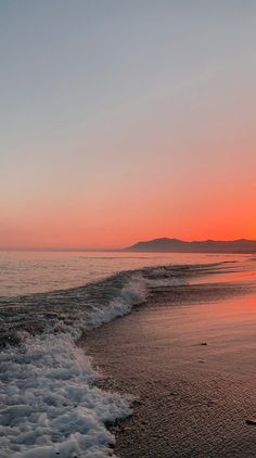 the sun is setting over the ocean with waves crashing on the shore and mountains in the distance