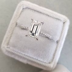 an emerald cut diamond ring in a white velvet box with diamonds on the band and side stones