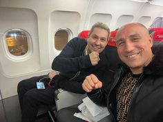 two men sitting on an airplane, one pointing at the camera and the other smiling