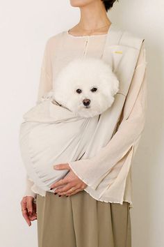 a woman is holding a white dog wrapped in a wrap on her back and wearing a beige top