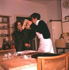 two people standing at a kitchen table with food in front of them and one person holding something up to his face