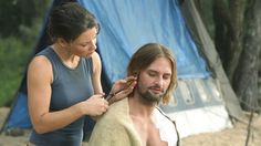 a man getting his hair cut by a woman in front of a blue tarp