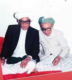 two men sitting next to each other in front of a white wall and red carpet