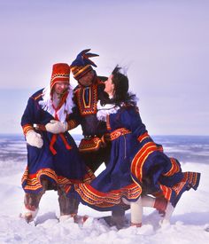 three people dressed in colorful clothing standing on snow covered ground with one person touching the other's ear