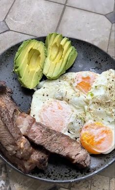 an egg, steak and avocado are on a black plate with a stone floor