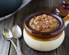 a wooden table with spoons and a bowl of food on it, next to a pot of stew