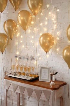 a table topped with lots of gold balloons and champagne glasses next to a white brick wall