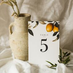 a table number card next to a vase with an orange and rosemary branch in it