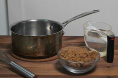 a wooden table topped with a metal pan and bowl filled with brown stuff next to a whisk