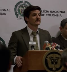 a man standing at a podium with microphones in front of him and two other men behind him