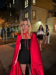 a woman in a red cape and black dress posing for the camera on a city street