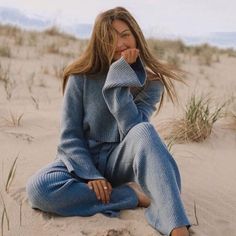 a woman sitting in the sand with her hand on her chin and looking off into the distance