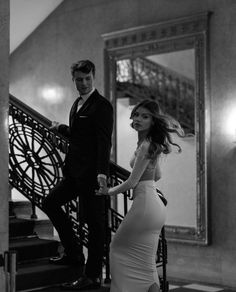 a woman in a white dress is walking down the stairs while a man in a tuxedo stands next to her