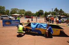 two men working on a car in the middle of a parking lot with people standing around