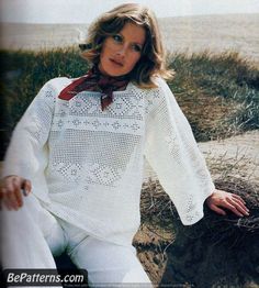 a woman sitting on top of a sandy beach next to grass and sand dunes wearing white clothing