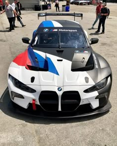 a bmw race car is parked in the parking lot with other cars behind it and people walking around