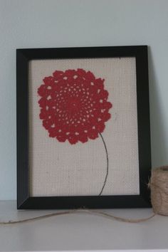 a red flower in a black frame sitting on a shelf next to twine rope